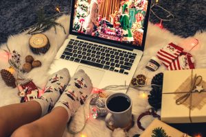 A person in snowman socks surrounded by holiday-themed trinkets watches a Christmas movie on their laptop
