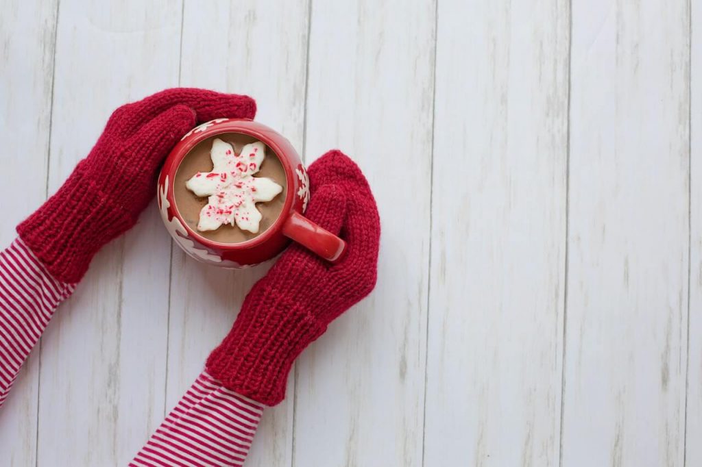 A person with red mittens on holds a cup of hot cocoa with two hands.