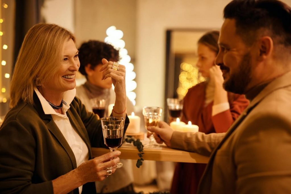 A woman and man, both holding drinks, converse with and smile at one another at a holiday party.